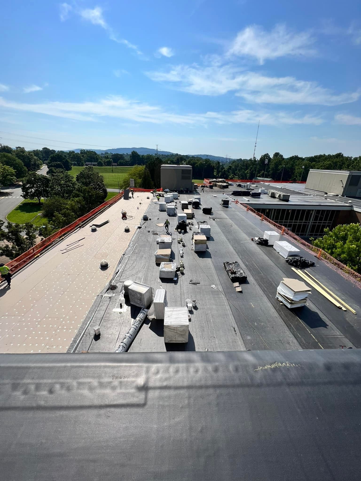 Commercial flat roof replacement in progress in Martinsburg WV with tan insulation board laid over the deck, dark single ply membrane rolled out, staged material pallets, and crew members in high visibility gear behind orange safety fencing