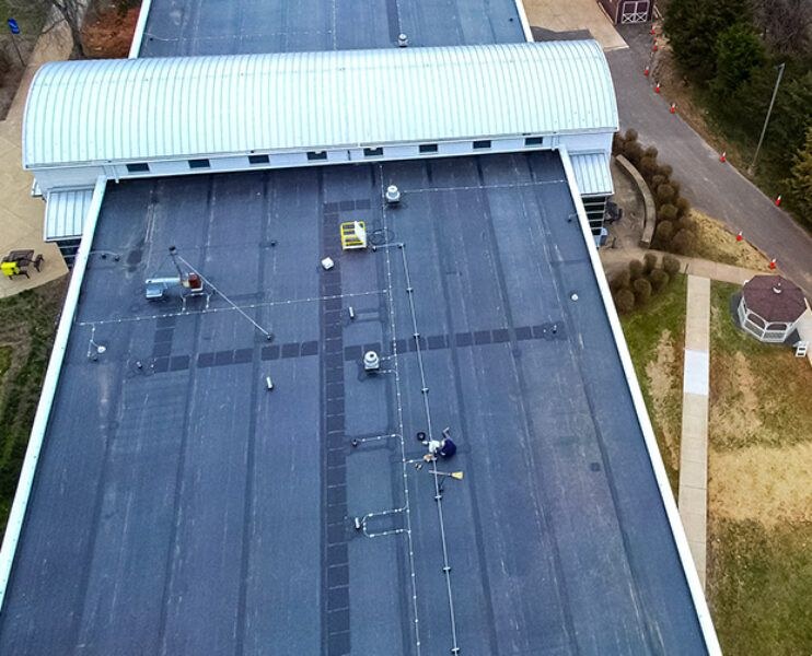 Overhead view of a worker on a dark low slope commercial membrane roof with fall protection lines rigged and a curved metal barrel roof section nearby, commercial roofing work by Black Stone Roofing of Martinsburg WV